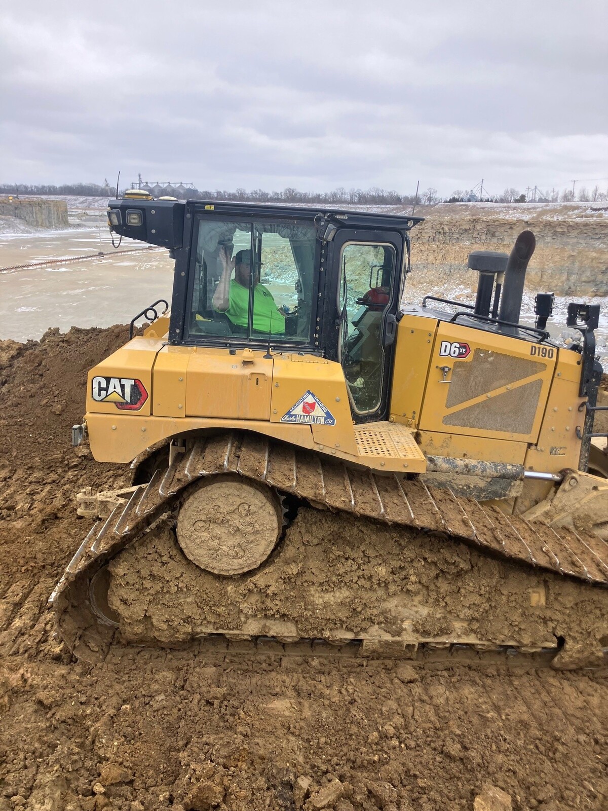 Construction worker operating a CAT D6 XE bulldozer on a muddy job site near a quarry under an overcast sky.